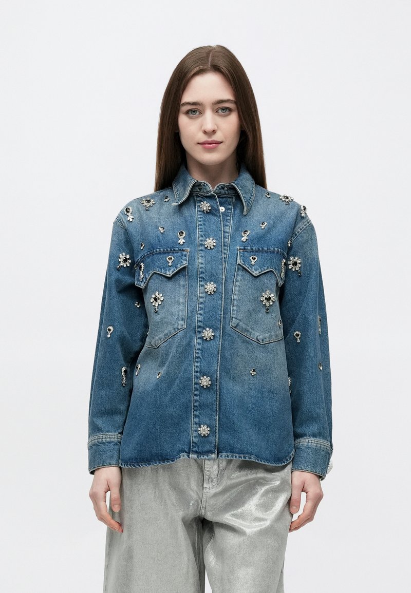 Νέα γυναίκα που φοράει μπλε τζιν μπουφάν με διακοσμητικά από λουλουδένιες στρας και ασημένιο μεταλλικό παντελόνι, στέκεται μπροστά σε ένα απλό λευκό φόντο.