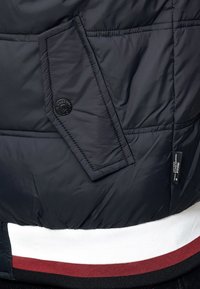 Granatowa kurtka puchowa z przekątną kieszenią zapinaną na czarny guzik, żebrowanym dołem w białe i burgundowe paski oraz z materiałową metką.