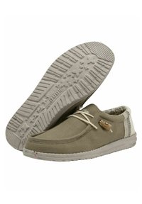 Zapatos casuales tipo slip-on, parte superior de tela verde oliva, suela de goma blanca con patrones de tracción en forma de estrella, detalles en tonos neutros y cordones decorativos.