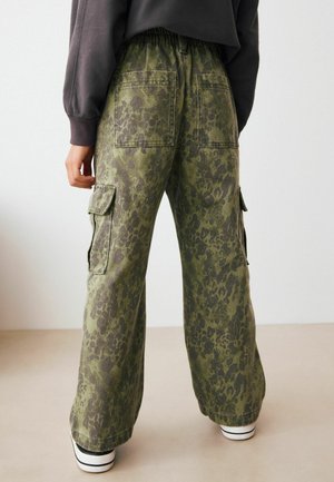 Person som bär löst sittande gröna camouflage cargo-byxor med bak- och sidofickor, i kombination med vita sneakers och en mörk långärmad topp.