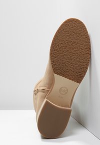 Bottine beige en daim avec une semelle en caoutchouc marron texturée et un logo discret au fond. Couture de la couture latérale visible.