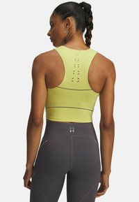 Žltý športový crop top s dizajnom racerback, ktorý má ventilačné otvory a kontrastné šedé akcenty. Skombinovaný s tmavošedými legínami s vysokým pásom.