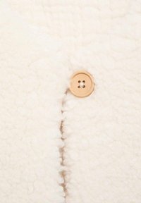 Țesătură albă cu o textură pufoasă și moale. Prezintă un nasture rotund de culoare bej deschis cu patru orificii, poziționat la cusătură.