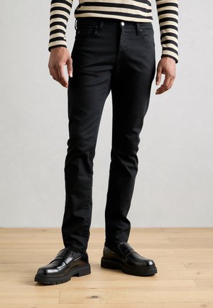 Personne portant un jean noir slim, des mocassins en cuir noir et une chemise à manches longues beige et noire rayée, debout sur un sol en bois.
