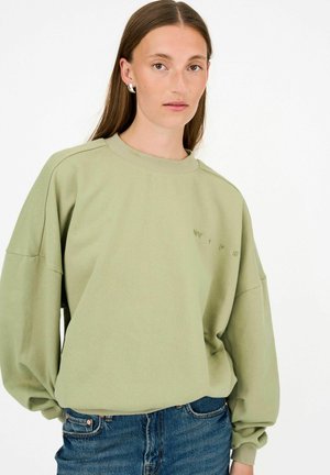 Donna con capelli castani lunghi che indossa una felpa oversize verde chiaro e jeans blu su uno sfondo bianco semplice.