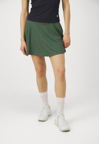 Zöld sport szoknya, fodros fazonban, sima anyagból készült. Fekete toppal és fehér sneakers-zel, bordázott fehér zoknikkal párosítva.