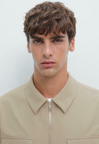 Beige zip-up jas met een kraag, zachte textuur, twee borstzakken, gedragen over een wit T-shirt. Close-up van het gezicht van een mannelijke model.