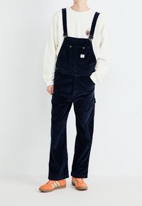 Navy Cord-Overall mit verstellbaren Trägern und großen Fronttaschen, kombiniert mit einem cremefarbenen Sweatshirt und orangefarbenen Turnschuhen mit blauen Streifen.