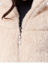 Zbliżenie na częściowo zapiętą beżową kurtkę sherpa na czarnej bluzce, ukazujące miękki, strukturalny materiał i srebrny suwak.