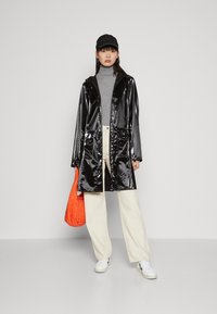 Svart glansig regnkappa med huva, framfickor, bärs över en grå polotröja och krämfärgade vida byxor, kombinerad med vita sneakers och en orange väska.