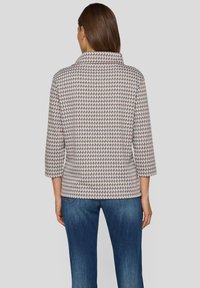 Frau in einer gemusterten Jacke mit 3/4-Ärmeln und blauen Jeans, die mit dem Rücken zu einem schlichten hellgrauen Hintergrund steht.