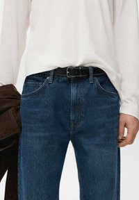 Pantalones vaqueros de mezclilla azul oscuro con corte recto, cinco bolsillos y costuras naranjas. Combinados con una camisa blanca de manga larga y un cinturón negro.