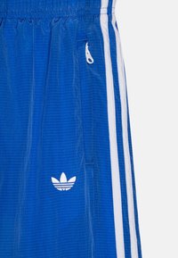 Modré sportovní šortky z texturované látky, s bílými bočními pruhy a bílým vyšitým logem. Elastický pas s detailním kapsou na zip.