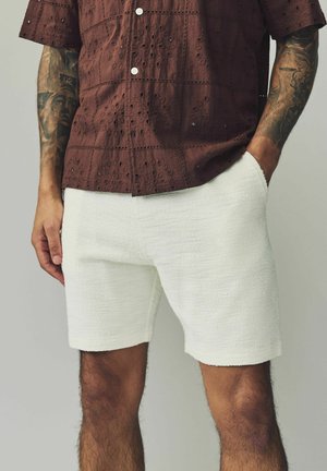White textured shorts with a relaxed fit, made from a woven material. Features side pockets and a clean hem. Paired with a brown patterned shirt.