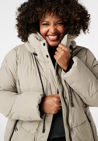 Manteau en duvet beige avec un col haut, un cordon de serrage et des poches zippées. Présente un matelassage horizontal et des éléments en métal noir pour le maintien.