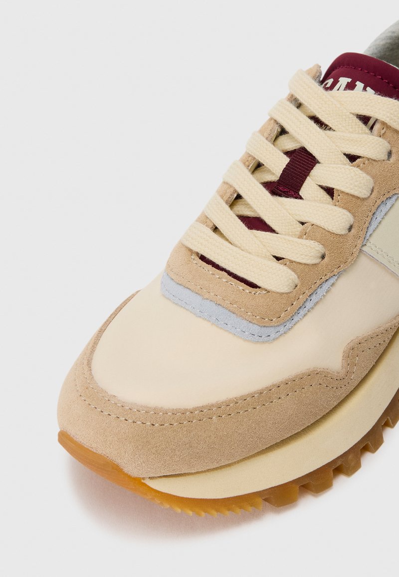 Beige und braune Wildledersneaker mit cremefarbenen Schnürsenkeln, burgunderfarbener Zunge und Details der Gummisohle auf weißem Hintergrund.