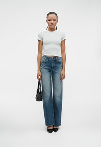 Λευκό τοπ με κοντά μανίκια, ψηλόμεσα μπλε τζιν με ξεθωριασμένο πλύσιμο και μαύρες γόβες με μυτερή μύτη. Κρατά μια μικρή μαύρη τσάντα.