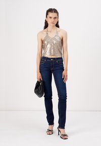 Metalni zlati crop top s sequini, temno modri skinny jeans, črna torbica, vzorčasti sandali na peto in nevtralna ozadja.