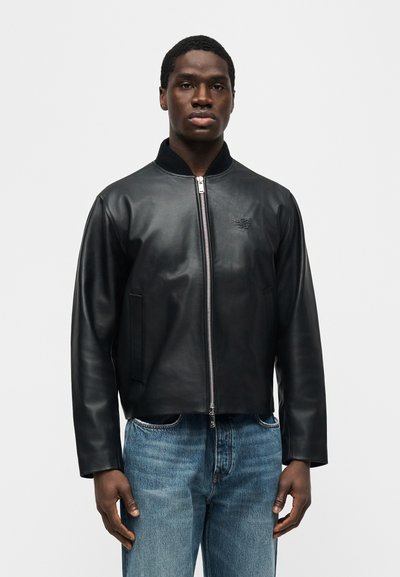 Νεαρός άνδρας στέκεται με το πρόσωπο προς τα εμπρός, φορώντας μαύρο δερμάτινο μπουφάν με φερμουάρ και μπλε τζιν, μπροστά σε απλό ανοιχτό γκρι φόντο.