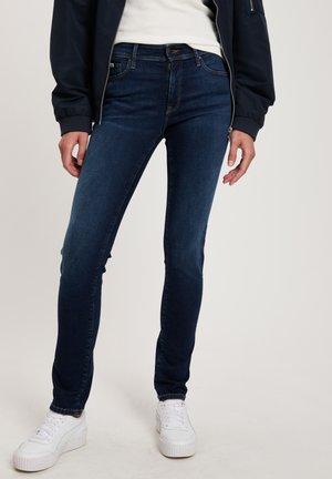 Skinny jeans in dark blue denim, featuring a mid-rise waist, classic 5-pocket design, subtle stitching, and worn with white sneakers.