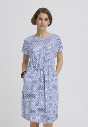 Žena s krátkymi hnedými vlasmi, oblečená v modro-bielych vzorovaných šatách dĺžky po kolená, s krátkymi rukávmi a vreckami, stojaca pred jednoduchým pozadím.