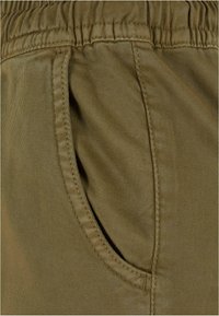 Gros plan sur une poche de pantalon vert olive avec des coutures visibles et une taille élastique au-dessus de l’ouverture de la poche.