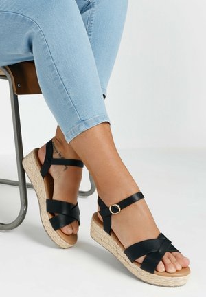 Zwarte wedge sandalen met kruisende banden, met een geweven espadrille zool en een gouden gespaccent. Op de achtergrond licht denim en een stoel.