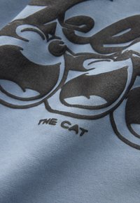 Jasnoniebieska tkanina z czarnym graficznym wzorem kota o przesadnych wyrazach twarzy. Zawiera napis "THE CAT".