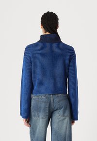 Blå ribbestrikket genser med høy krage og lange ermer, med kontrasterende mørke striper, kombinert med lyse denimbukser.