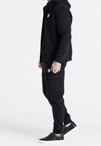 Černá mikina s kapucí a tepláky z bavlněné směsi, vybavené zipem, bočními kapsami a vyšívaným logem, doplněné černými teniskami.