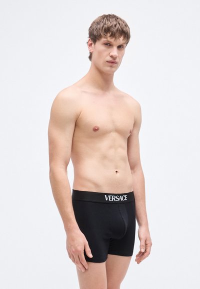 Boxer en coton noir avec une ceinture élastique arborant le logo "VERSACE" en blanc. Le modèle est ajusté avec une texture lisse.
