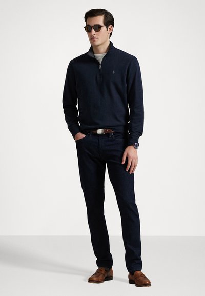 Marineblå zip-up genser laget av strikket stoff, mørke jeans, brune lærsko og svarte solbriller; modellen står med hendene i lommene.