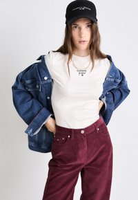 Τζάκετ από τζιν με φαρδιά μανίκια, κρέμα μπλούζα με λογότυπο, μπορντώ παντελόνι από κοτλέ, μαύρο καπέλο με κείμενο μάρκας, και κολιέ από πέρλες.