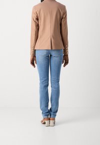 Blazer en similicuir beige au design ajusté, avec des boutons décoratifs sur les manches, assorti à un jean bleu clair et des sandales à talons beiges.