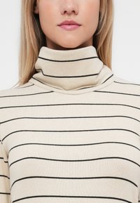 Beige garbónyakú pulóver fekete vízszintes csíkokkal, puha anyagból készült, magas gallérral és laza szabással. Simaságával jellegzetes minták nélkül.