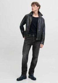 Chaqueta de cuero negra con un cuello alto, combinada con una camiseta negra acanalada y vaqueros negros descoloridos. El modelo lleva botas oscuras y un cinturón con detalles metálicos.