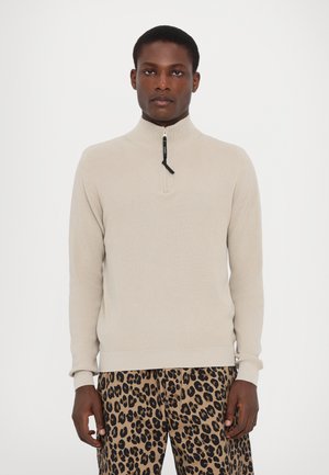 Pulover bej din tricotaj cu guler înalt și închidere cu fermoar pe jumătate. Dispune de o textură cu nervuri și mansete ajustate, asortat cu pantaloni cu imprimeu de leopard.