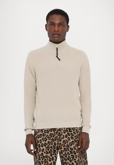 Beige knit sweater with a high collar and half-zip closure. Features ribbed texture and fitted cuffs, paired with patterned leopard-print pants.