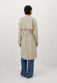 Beige trench coat with a belted waist, double-breasted front, and back flap detail, paired with blue wide-leg jeans. Smooth fabric texture.