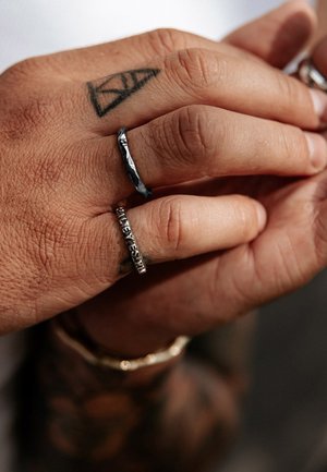 Two rings on a hand: a textured black band and a silver band with engraved letters. The hand has visible skin texture and a tattoo.