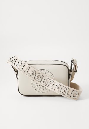 Beige crossbody tas met een geperforeerd cirkelontwerp, zwarte afwerking en een afneembare band met de letters "KARL LAGERFELD" in een metallic afwerking.