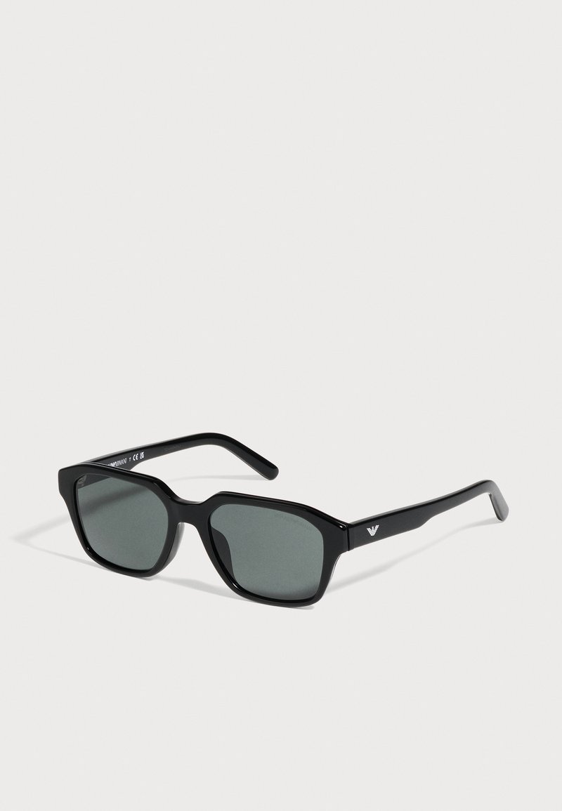 Lunettes de soleil noires rectangulaires, avec des verres gris foncé et un petit logo blanc sur la branche, sur fond clair.