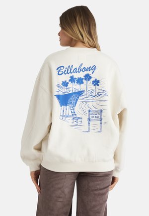 Kobieta w białej bluzie z niebieskim widokiem plaży, palmami, chatką i tekstem "Billabong" na plecach.