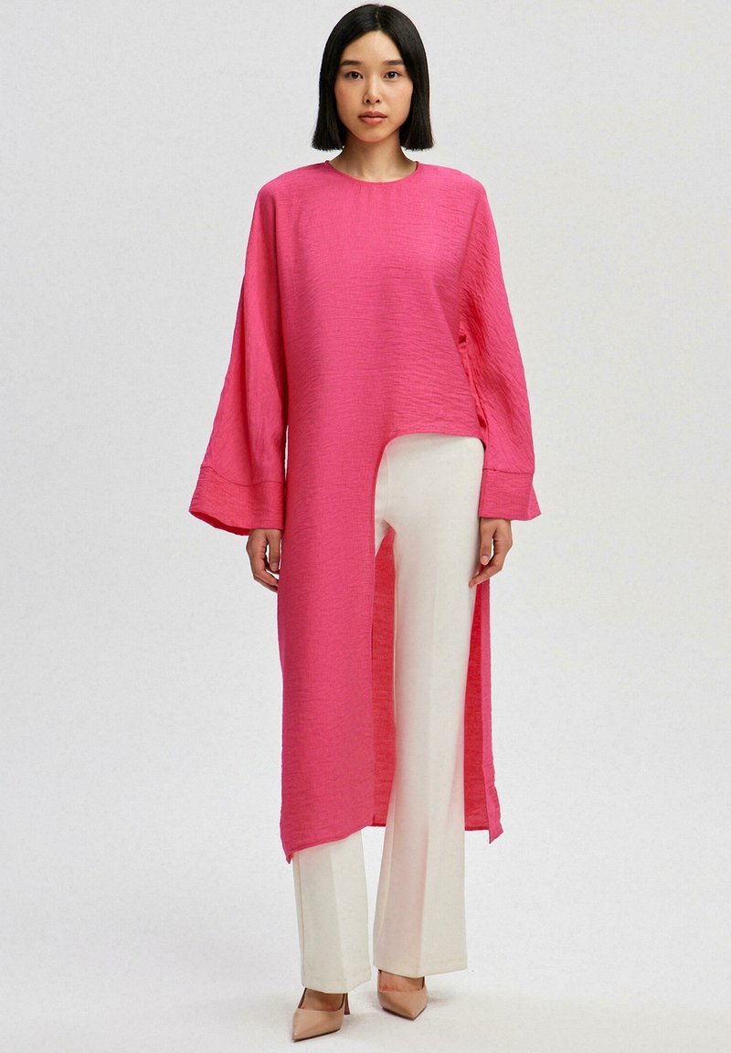 Túnica larga rosa con tejido texturizado, mangas amplias y dobladillo asimétrico, combinada con pantalones blancos y tacones nude. Diseño minimalista.