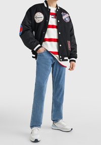 Černá varsity bunda s bílými a červenými nášivkami, pod ní pruhované červenobílé tričko, světle modré džíny a bílé tenisky.