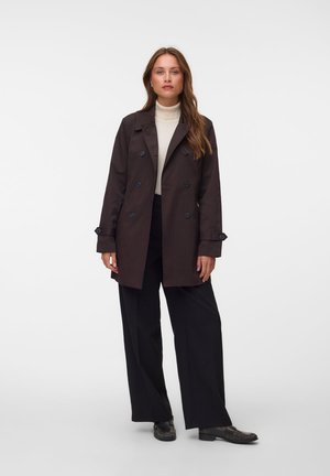 Une femme porte un manteau marron foncé boutonné sur un col roulé crème, un pantalon noir ample et des chaussures noires, devant un fond de studio uni.