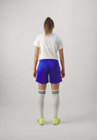 Balts T-kreklis, zili šorti ar oranžiem akcentiem, ceļa auguma balti zeķes ar melnām svītrām un spilgti dzelteni futbola apavi.