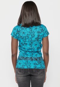 T-shirt turquoise à manches courtes avec un motif floral noir, design ajusté et texture subtile. Associé à un jean foncé.