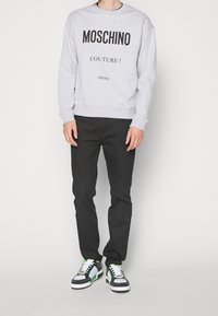 Šedá mikina s kulatým výstřihem a černým nápisem "MOSCHINO COUTURE! Milano", doplněná o černé kalhoty a víceméně barevné tenisky.