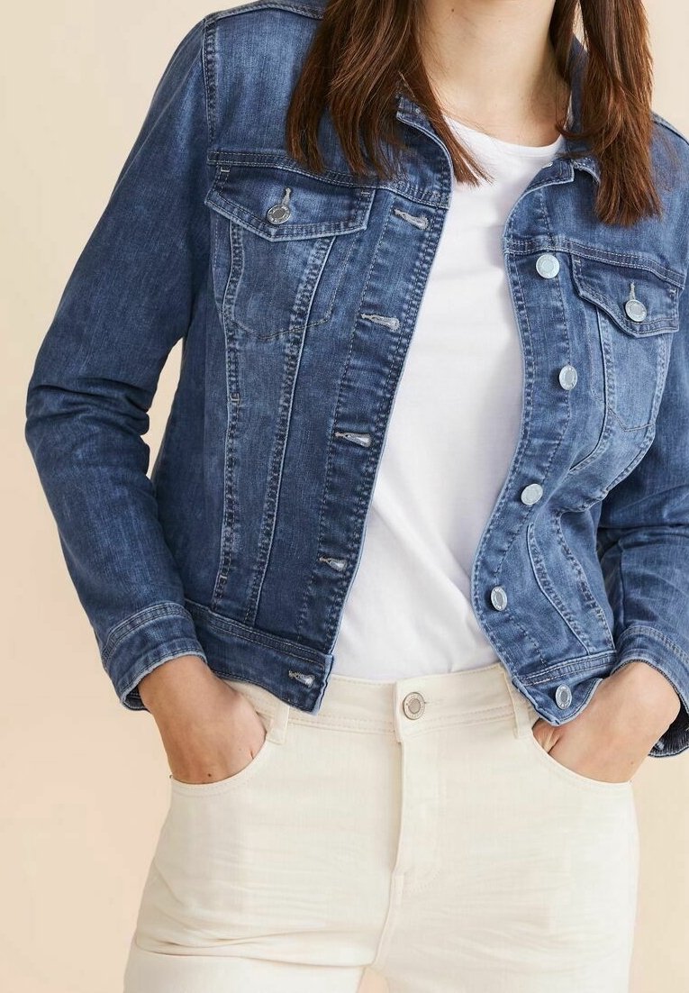 Femme portant une veste en denim bleu sur un t-shirt blanc, les mains dans les poches d'un pantalon beige clair, devant un fond uni.